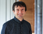 Eine Person mit kurzen dunklen Haaren lächelt und trägt ein dunkles Hemd mit Knöpfen. Der Hintergrund, der an die Architektur der Hochschule Coburg erinnert, besteht aus vertikalen Linien und Säulen, die der Szene eine akademische Eleganz verleihen.