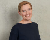 Eine lächelnde Person mit zurückgebundenem hellbraunem Haar und einem dunklen Hemd steht vor einem strukturierten, hellgrauen Hintergrund an der Hochschule Coburg.