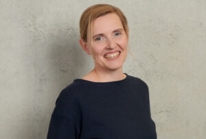 A person with short blonde hair smiles at the camera, wearing a dark blue top. They stand against a plain, light-colored textured wall, evoking the serene atmosphere of Hochschule Coburg's campus, creating a simple and relaxed portrait.