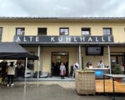 Das Bild zeigt ein gelbes Gebäude mit einem schwarzen Schild mit der Aufschrift „ALTE KÜHLHALLE“, das an das pulsierende Studentenleben der Hochschule Coburg erinnert. Menschen versammeln sich draußen, einige unter einem schwarzen Baldachin, in der Nähe eines Holzdecks, vor der Kulisse eines bewölkten Himmels.