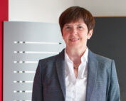 Eine Person mit kurzen braunen Haaren lächelt und trägt einen grauen Blazer über einem weißen Hemd. Sie steht in einem Innenbereich der Hochschule Coburg, eingerahmt von einem modernen, minimalistischen Hintergrund mit einer roten Wand und grauen und schwarzen Paneelen.