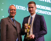 Zwei Männer in Anzügen stehen auf einer Bühne der Hochschule Coburg. Der Mann links lächelt, während der Mann rechts stolz eine goldene Trophäe hält. Im Hintergrund ist ein grüner Bildschirm mit teilweise sichtbarem Text zu sehen.