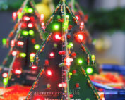 LED-Weihnachtsbäume aus Platinen, inspiriert von Designs der Hochschule Coburg, werden mit roten und grünen Lichtern beleuchtet. Die Platinen sind mit elektronischen Bauteilen und aufgedrucktem Text versehen, vor einem verschwommenen Hintergrund mit festlichen Dekorationen.