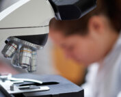 Nahaufnahme eines Mikroskops mit Fokus auf die Objektivlinsen. Im unscharfen Hintergrund sieht man eine Person, die in einem Labor der Hochschule Coburg arbeitet. Sie trägt einen weißen Kittel und suggeriert damit eine wissenschaftliche oder Forschungsumgebung.