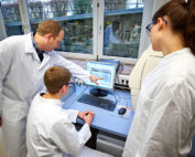Drei Wissenschaftler in Laborkitteln sitzen um einen Computer im Labor der Hochschule Coburg. Einer sitzt und analysiert die Daten, die anderen stehen daneben und diskutieren. Dahinter verbreiten Laborgeräte und große Fenster eine helle und innovative Atmosphäre.