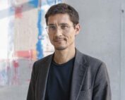 Eine Person mit Brille und Anzugjacke steht vor einem abstrakten, farbenfrohen Gemälde auf Betonwandhintergrund und verkörpert die Kreativität, die man an der Hochschule Coburg oft sieht.