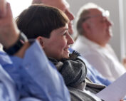 Eine Person mit kurzen Haaren und Kopftuch sitzt lächelnd in einer Reihe von Menschen bei einer Indoor-Veranstaltung der Hochschule Coburg. Sie hält ein Blatt Papier in der Hand und auch andere neben ihr sind engagiert. Der Fokus liegt auf dem fröhlichen Ausdruck.