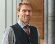 Ein Mann mit kurzen hellbraunen Haaren und Brille, der ein gepunktetes Hemd, eine rote Krawatte und eine schwarze Weste trägt, steht in der modernen Innenarchitektur der Hochschule Coburg, umgeben von vertikalen Holz- und Metallpaneelen.