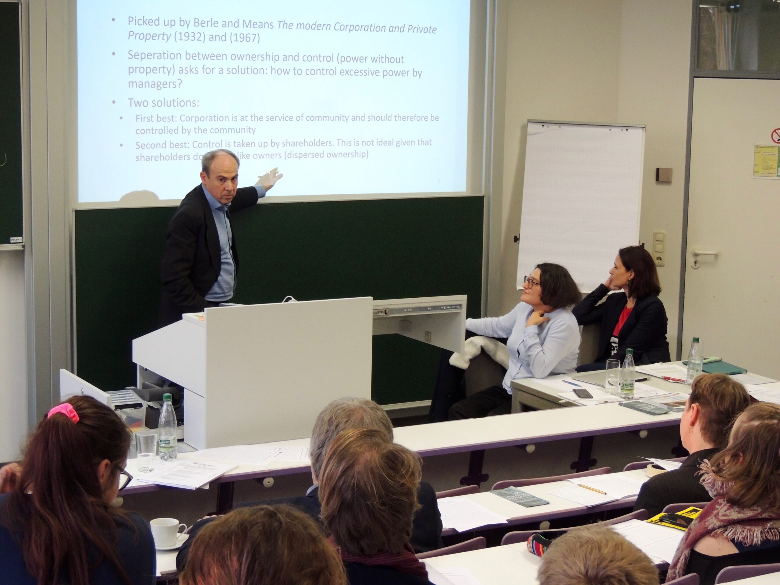 Ein Redner der Hochschule Coburg zeigt auf eine Präsentationsfolie in einem Klassenzimmer, während ein erwachsenes Publikum aufmerksam zuhört. Der Raum ist mit Tischreihen gesäumt, und eine Person macht sich fleißig Notizen. Im Hintergrund sind eine Tafel und ein großes Whiteboard zu sehen.