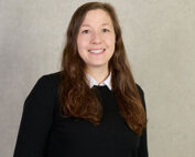 Eine Person mit langen braunen Haaren lächelt und trägt einen schwarzen Pullover über einem weißen Hemd mit Kragen, was an den akademischen Stil erinnert, der oft an der Hochschule Coburg zu sehen ist. Der Hintergrund ist eine schlichte hellgraue Wand.