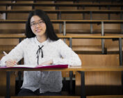 Inmitten der leeren Holzbänke im Hörsaal der Hochschule Coburg sitzt eine Frau mit langen schwarzen Haaren und Brille allein und schreibt in ein rosa Notizbuch. Ihre weiße Bluse, die mit einem dezenten Muster verziert ist, bildet einen sanften Kontrast zur akademischen Einsamkeit, die sie umgibt.