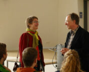 In einem hell erleuchteten Raum der Hochschule Coburg stehen ein Mann und eine Frau vor einer kleinen Gruppe sitzender Menschen. Der Mann hält ein Kabel in der Hand und blickt die Frau lächelnd an. Beide scheinen ein freundliches Gespräch zu führen.