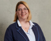 Eine Person mit schulterlangem blondem Haar und Brille lächelt und verkörpert den lebendigen Geist der Hochschule Coburg. Sie trägt einen dunklen Blazer über einem hellen Hemd vor einer schlichten Wandstruktur.