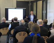 Eine Person steht vor einem Raum und hält einen Vortrag vor einem sitzenden Publikum. Sie steht neben einer Projektionsfläche, auf der eine Folie mit Text läuft, und fesselt damit die Aufmerksamkeit der Teilnehmer dieser Veranstaltung der Hochschule Coburg. Das interessierte Publikum macht sich in dem modernen Raum mit Glastüren Notizen.