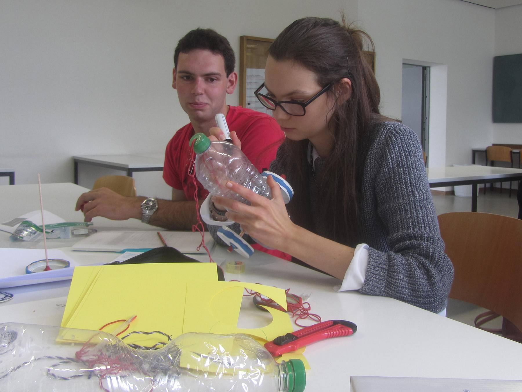 Zwei Personen sitzen an einem Tisch in einem Hörsaal der Hochschule Coburg und sind in ihr Projekt vertieft. Die Frau hält eine Plastikflasche und trägt Klebstoff auf, während der Mann zusieht. Auf dem Tisch liegen verstreut Materialien wie Papier, Werkzeuge und Drähte, was die Kreativität widerspiegelt, die an dieser Institution gefördert wird.