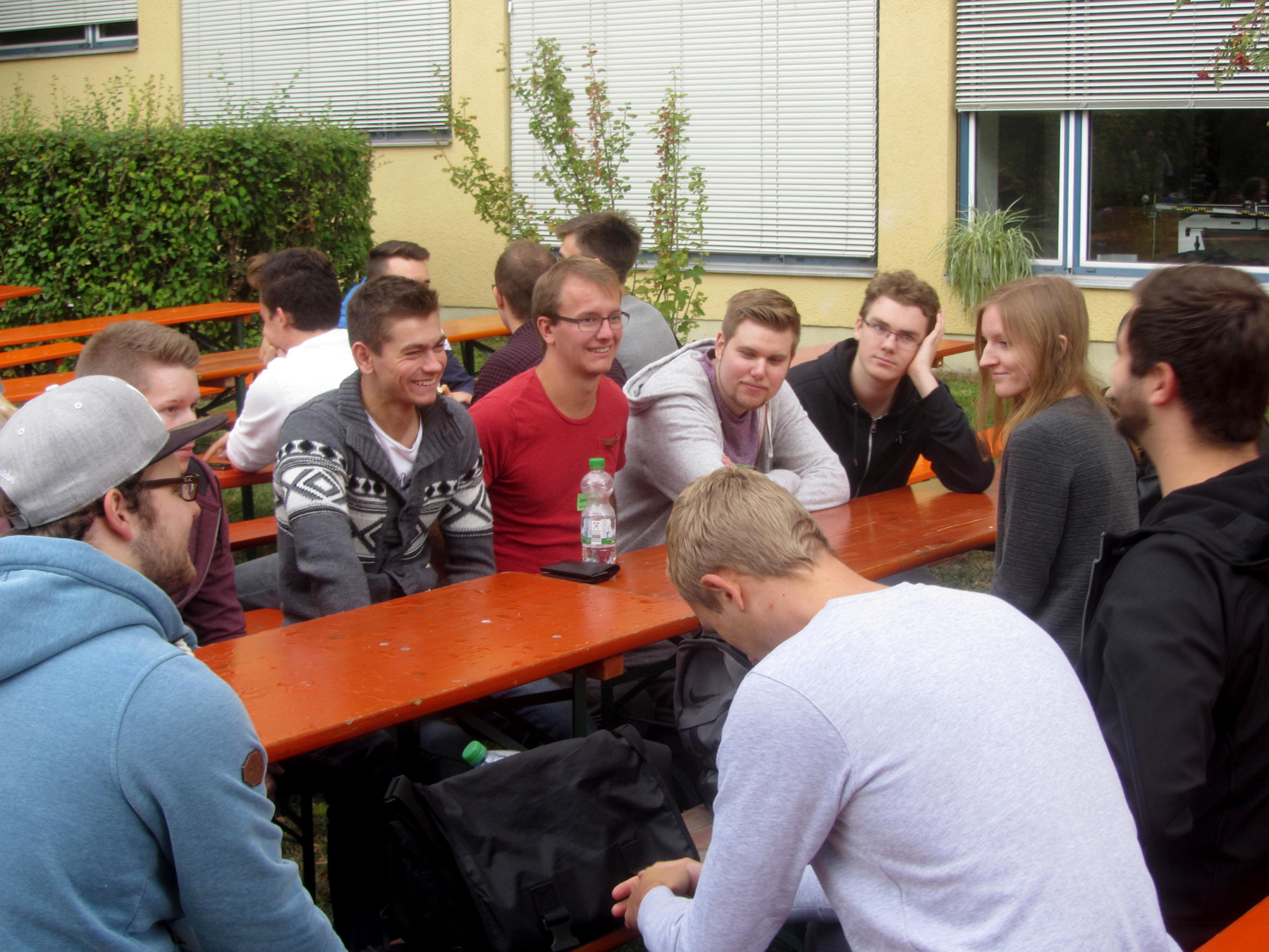 Eine Gruppe von Menschen, die überwiegend legere Kleidung wie Pullover und Jacken tragen, sitzt um eine Holzbank im Außenbereich der Hochschule Coburg. Sie unterhalten sich und scheinen die Gesellschaft der anderen an diesem schönen Tag zu genießen.