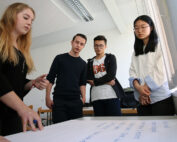 Vier Personen stehen an einem Tisch in der Hochschule Coburg und diskutieren. Eine Frau deutet auf ein großes Blatt Papier mit Notizen, die anderen beobachten aufmerksam. Sie befinden sich in einem gut beleuchteten Raum mit weißen Wänden, was eine Atmosphäre konzentrierter Zusammenarbeit fördert.