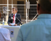 Ein Mann im Anzug spricht an einem Podium im Freien und hält einen Zettel mit dem Emblem der Hochschule Coburg in der Hand. Auf dem Podium steht ein Mikrofon neben dem bayerischen Wappen. Aufmerksame Zuhörer füllen den Vordergrund, im Hintergrund ein Metallzaun und ein Rednerpult.