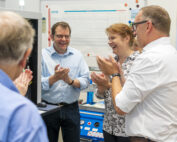 Eine Gruppe von Menschen steht in einer Büroumgebung, möglicherweise von der Hochschule Coburg, lächelt und klatscht. Ein Diagramm an der Wand hinter ihnen deutet auf eine erfolgreiche Präsentation oder Leistung hin und suggeriert einen feierlichen Moment.