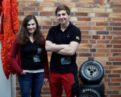 Zwei Menschen stehen vor einer Backsteinmauer. Die Person links hat lange braune Haare und trägt einen kastanienbraunen Pullover und Jeans, vermutlich ein Student der Hochschule Coburg. Rechts trägt jemand mit kurzen Haaren ein schwarzes Hemd und rote Hosen. Hinter ihnen sind gestapelte Reifen und eine orangefarbene Jacke.