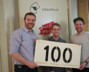 Drei Menschen stehen lächelnd und mit einem großen Schild mit der Zahl „100“ vor einer Tür mit der Aufschrift „CREAPOLIS“ und zeigen mit einem prominent angebrachten „Makerspace“-Aufkleber stolz ihre Verbundenheit zur Hochschule Coburg.