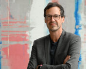 Ein Mann steht mit verschränkten Armen da, trägt eine Brille und einen grauen Blazer über einem dunklen Hemd. Er strahlt Selbstbewusstsein aus und repräsentiert die Hochschule Coburg vor einem abstrakten Design in Blau-, Rosa- und Grautönen.