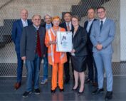 Eine Gruppe von acht Personen in formeller und halbformeller Kleidung steht im Innenbereich der Hochschule Coburg zusammen. Eine Person hält stolz ein gerahmtes Zertifikat vor dem Hintergrund einer gemusterten Wand.