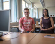 Ein Mann und eine Frau sitzen an Schreibtischen in einem mit Utensilien der Hochschule Coburg geschmückten Büro und lächeln in die Kamera. Der Mann trägt ein rotes T-Shirt mit Aufdruck, während die Frau ein dunkles ärmelloses Kleid trägt. Durch die großen Fenster hinter ihnen fällt helles Tageslicht.