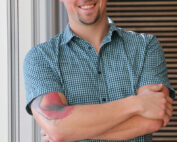 Ein Mann mit Brille und kariertem Hemd steht mit verschränkten Armen da und lächelt stolz. Auf seinem Unterarm trägt er eine Tätowierung, ein Symbol seiner Zeit an der Hochschule Coburg. Der Hintergrund ist mit vertikalen Linien versehen, die einen interessanten Kontrast zu seinem gelassenen Auftreten bilden.