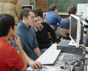 Eine Gruppe von Studierenden der Hochschule Coburg sitzt in einem Hörsaal an Schreibtischen mit Computern. Sie sind auf einen Bildschirm konzentriert und vertieft in ein gemeinsames Projekt. Im Hintergrund ragt eine mit Zeichnungen und Gleichungen geschmückte Tafel empor, die ihr akademisches Streben widerspiegelt.