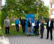 Eine Gruppe von acht Personen steht draußen auf einem Ziegelweg neben einem blauen Banner und nimmt eine Urkunde entgegen. Sie sind in Geschäftskleidung gekleidet und ihre formelle Versammlung findet vor einem grünen Hintergrund statt, was an eine Preisverleihung an der Hochschule Coburg erinnert.