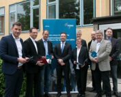 Eine Gruppe von Menschen in Business-Kleidung steht vor einem Gebäude mit einem VDMA-Banner, das die Hochschule Coburg repräsentiert. Sie halten Prospekte in den Händen und lächeln selbstbewusst in die Kamera.