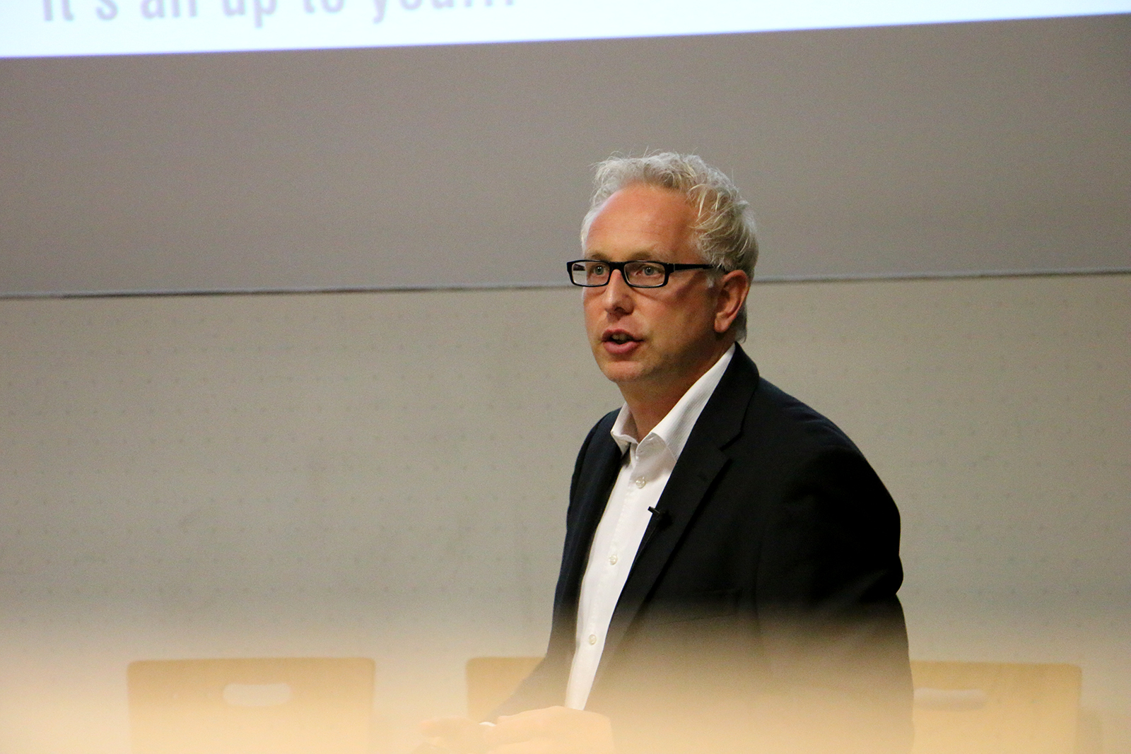 Ein Mann mit grauen Haaren und Brille spricht vor einem Publikum an der Hochschule Coburg. Er trägt einen schwarzen Anzug und ein weißes Hemd. Hinter ihm lässt eine Projektionsfläche mit teilweise sichtbarem Text auf einen spannenden Vortrag oder eine Präsentation schließen und weckt damit das Interesse der aufmerksamen Menge.