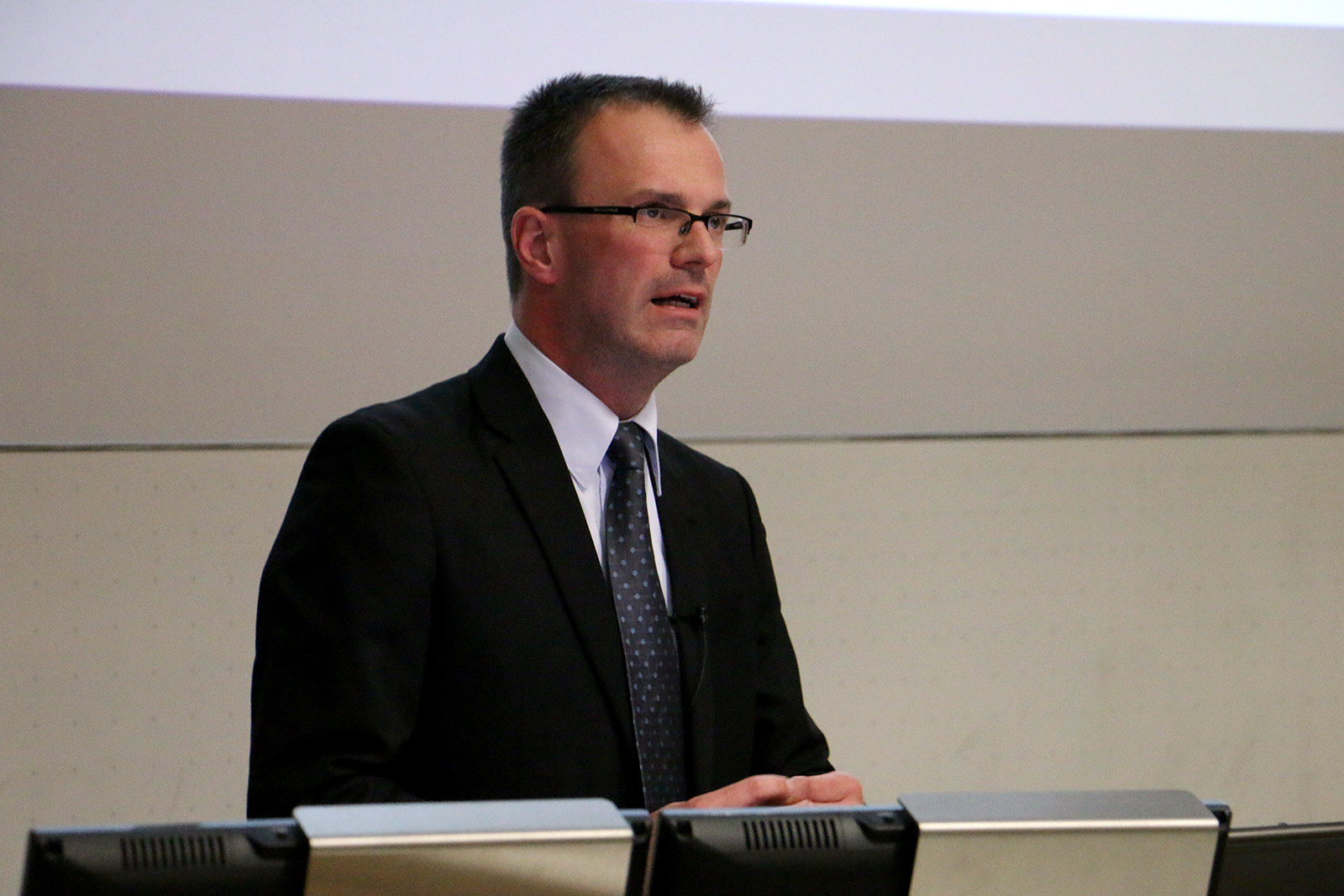 Ein Mann in Anzug und Krawatte spricht an einem Podium und vertritt die Hochschule Coburg. Im Hintergrund ist eine schlichte Wand mit einer Projektionsfläche zu sehen. Er trägt eine Brille und hat ein Ansteckmikrofon an seiner Jacke befestigt. Er ist bereit, selbstbewusst vor dem Publikum zu sprechen.