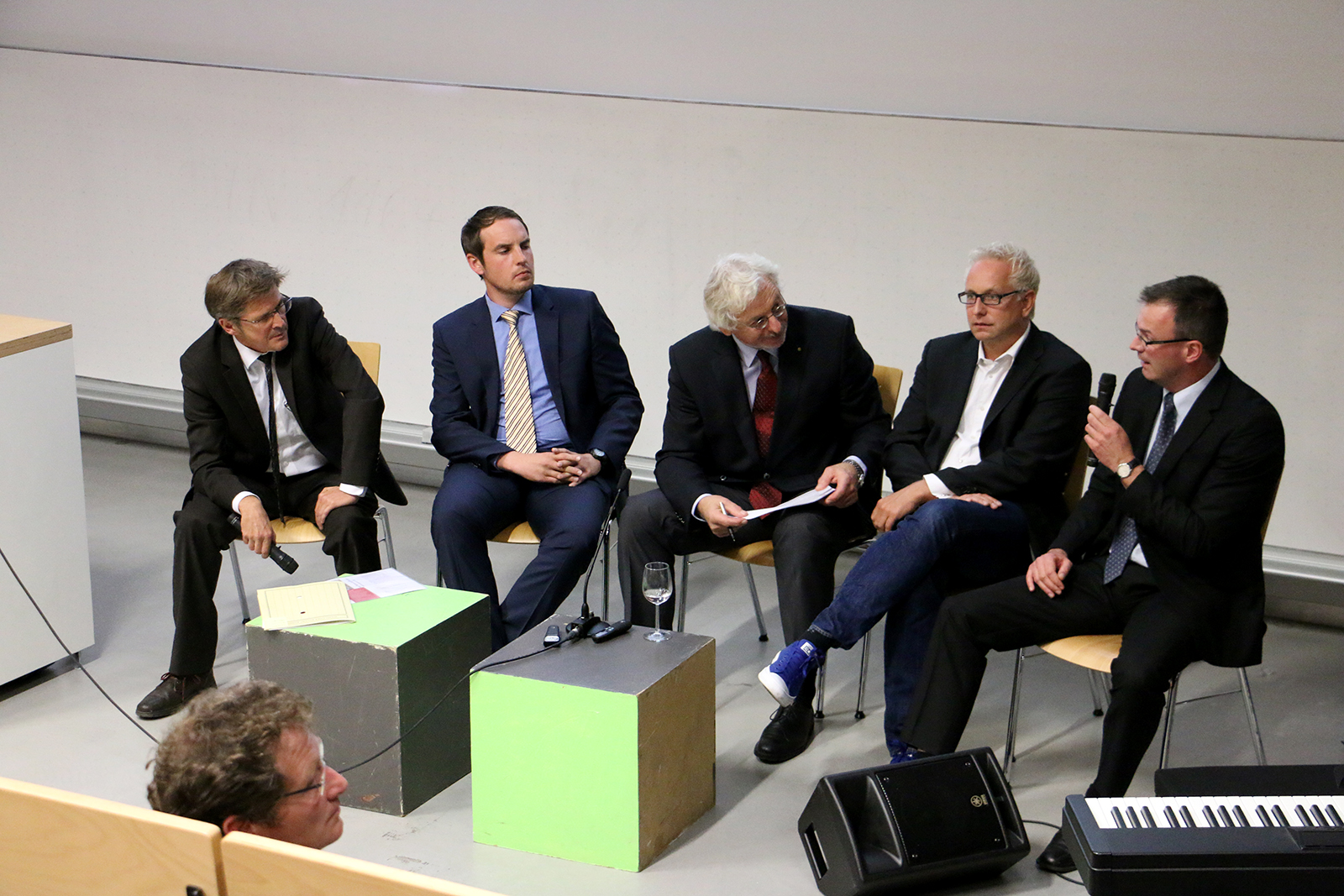 Eine Gruppe von fünf Männern in Anzügen sitzt in einem Konferenzraum der Hochschule Coburg, davor stehen zwei bunte Kisten. Ein Mann spricht in ein Mikrofon, während die anderen aufmerksam zuhören. Im Vordergrund ist eine Tastatur teilweise zu sehen.