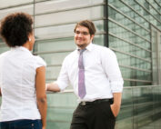 Zwei Menschen im Business-Outfit unterhalten sich im Freien nahe der modernen Glasfassade der Hochschule Coburg. Einer, in weißem Hemd und Krawatte, lehnt lässig an einem Geländer, die andere, in weißer Bluse, steht ihm gegenüber.