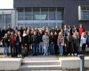 Eine große Gruppe von Menschen steht auf den Stufen vor einem modernen Gebäude der Hochschule Coburg. Sie sind leger und halbformell gekleidet, lächeln und posieren unter einem klaren Himmel für die Kamera.