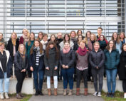 Eine Gruppe von Menschen posiert gemeinsam vor dem modernen Gebäude der Hochschule Coburg mit großen Fenstern. Sie tragen lässige Winterkleidung, darunter Mäntel und Schals. Das Wetter scheint kühl und bewölkt zu sein.