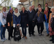 Eine Gruppe von Menschen steht zusammen auf einem Bürgersteig vor der Hochschule Coburg und posiert für ein Foto. Sie sind alle leger und warm gekleidet, und einige lächeln. Im Hintergrund sind Bäume und Gebäude zu sehen.