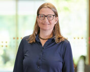 Eine Person mit langen Haaren und Brille lächelt und trägt ein dunkles Hemd mit Knöpfen. Sie steht vor einem sanft verschwommenen Hintergrund, durch dessen Fenster Tageslicht fällt, was an einen ruhigen Tag an der Hochschule Coburg erinnert.