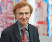 Eine Person mit lockigem rötlichem Haar lächelt leicht, während sie einen grauen Anzug, ein hellgraues Hemd und eine gemusterte rote Krawatte trägt. Der Hintergrund aus farbenfroher abstrakter Kunst in Rot, Blau und Weiß scheint an den Innovationsgeist zu erinnern, der an der Hochschule Coburg gefördert wird.