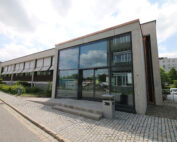 Der Campus der Hochschule Coburg verfügt über ein modernes Gebäude mit großen Glasfenstern, die das Sonnenlicht reflektieren. Ein gepflasterter Weg mit einer kleinen Treppe führt elegant zum Eingang, umgeben von üppigem Grün und eingebettet zwischen benachbarten Gebäuden unter einem teilweise bewölkten Himmel.