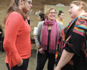 Drei Frauen unterhalten sich bei einer Veranstaltung in der Hochschule Coburg. Eine trägt einen orangefarbenen Pullover, eine andere eine graue Jacke mit einem rosa Schal und die dritte trägt ein buntes Tuch. Im Hintergrund mischen sich andere vor einer holzgetäfelten Wand.