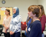 Eine bunt gemischte Gruppe steht in einem Raum der Hochschule Coburg und diskutiert angeregt. Eine Frau mit blauem Hijab und weitere leger gekleidete Personen blicken nach vorne, während eine Pinnwand mit Haftnotizen als Hintergrund dient.