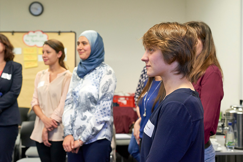 Eine bunt gemischte Gruppe steht in einem Raum der Hochschule Coburg und diskutiert angeregt. Eine Frau mit blauem Hijab und weitere leger gekleidete Personen blicken nach vorne, während eine Pinnwand mit Haftnotizen als Hintergrund dient.