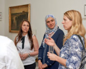 Bei einer Veranstaltung in der Hochschule Coburg unterhalten sich vier Frauen. Eine trägt ein geblümtes Top, eine andere ein Kopftuch und eine dritte eine weiße Bluse mit schwarzer Krawatte. An der Wand hinter ihnen ist ein gerahmtes Gemälde zu sehen. Sie halten Gläser in den Händen, was auf eine lebhafte gesellige Runde schließen lässt.