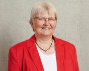 Eine Person mit kurzen, hellen Haaren lächelt und trägt eine Brille, einen roten Blazer und ein weißes Oberteil vor einem schlichten grauen Hintergrund und verkörpert den lebendigen Geist der Hochschule Coburg.