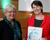 Zwei Frauen stehen drinnen und lächeln herzlich. Die Frau links trägt ein schwarzes Oberteil, während ihre Begleiterin im roten Blazer stolz ein Zertifikat der Hochschule Coburg aus dem Jahr 2015 in den Händen hält. Sie sind umgeben von holzgetäfelten Wänden und im Hintergrund leuchten sanft Kerzen.