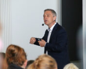 Ein Mann in dunklem Anzug und weißem Hemd spricht zu einem Publikum an der Hochschule Coburg. Er trägt ein Headset-Mikrofon. Er gestikuliert energisch mit den Händen, während im Vordergrund unscharfe Zuschauer zu sehen sind.