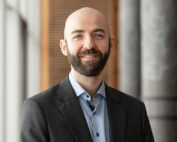 Ein kahlköpfiger Mann mit Bart, einem dunklen Jackett und einem hellblauen Hemd steht lächelnd im Innenbereich der Hochschule Coburg neben einem großen Fenster und einer Holzpaneel.